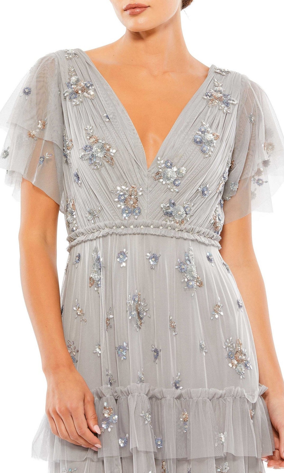 A woman wears the Mac Duggal 9068 Short Sleeve Platinum Formal Gown, featuring a light gray color, deep V-neckline, floral embroidery, and layered tulle details.